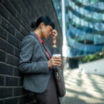 businesswoman leaning against a wall, holding a coffee, and rubbing her temple due to stress or headache