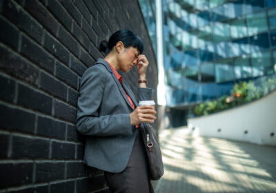 businesswoman leaning against a wall, holding a coffee, and rubbing her temple due to stress or headache