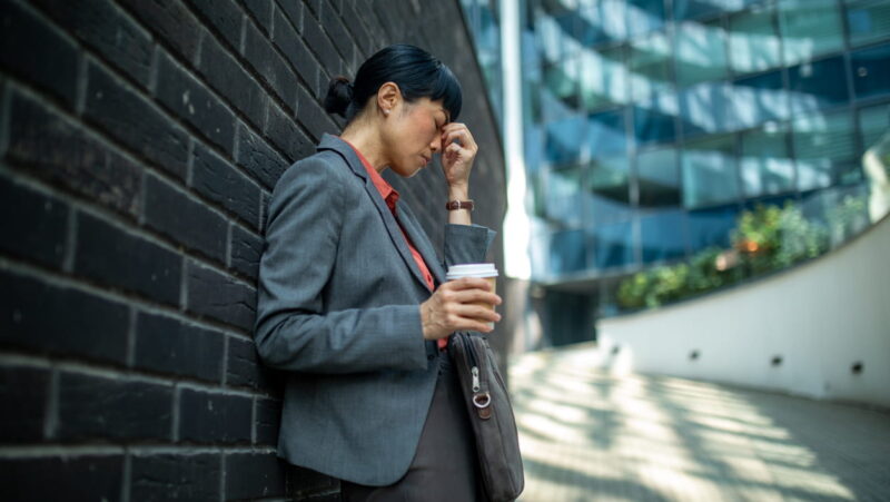 businesswoman leaning against a wall, holding a coffee, and rubbing her temple due to stress or headache