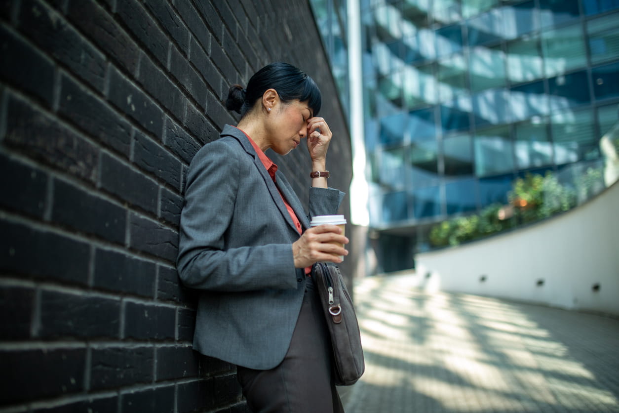businesswoman leaning against a wall, holding a coffee, and rubbing her temple due to stress or headache